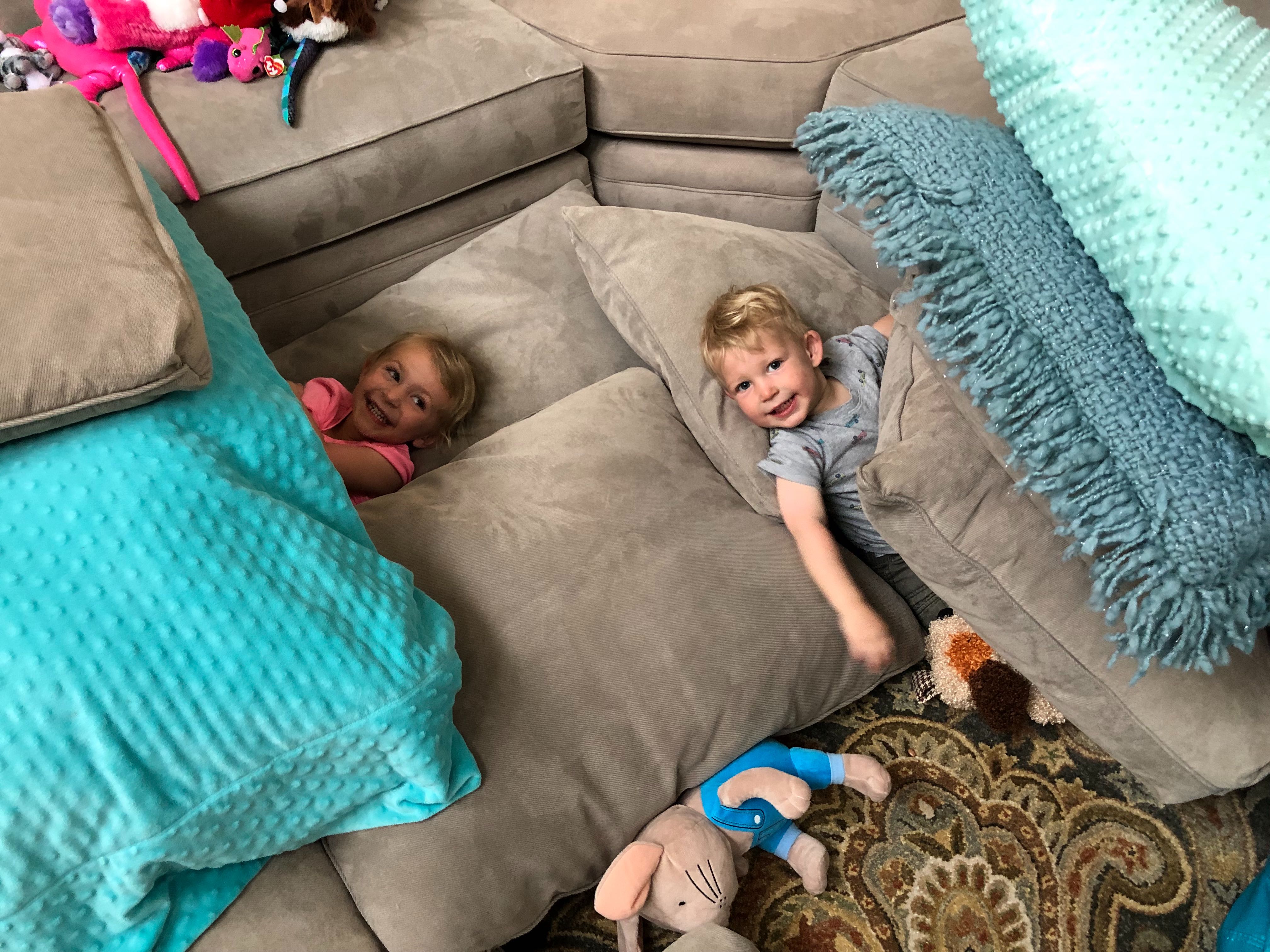 Two children hiding out under mattress and pillows with big smiles on their faces.