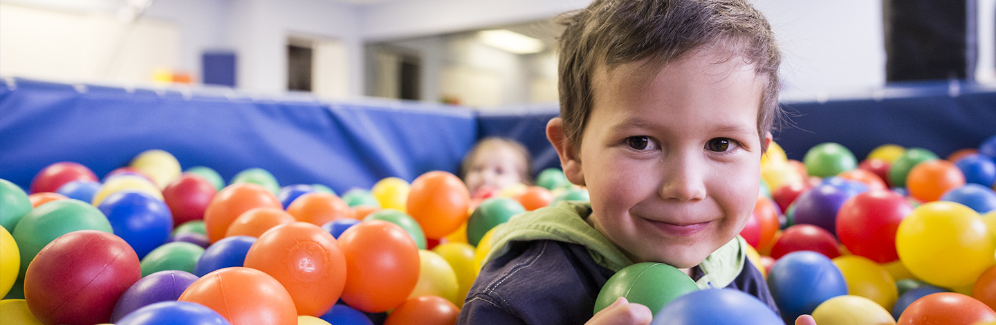 child in a ball bit, with colorful plastic balls up to his shoulders. He is looking at the camera with a smile.
