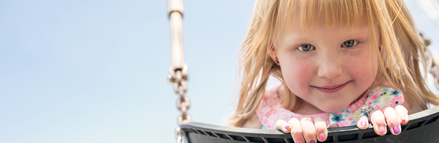 A young girl with strawberry blonde hair looking at the camera smiling while sitting in a swing