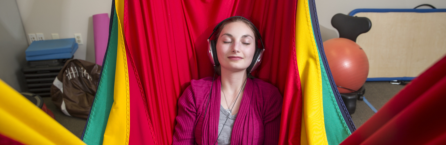 A young adult sitting in a colorful fabric hammock closing her eyes with headphones on