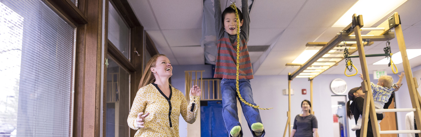 A child is hanging onto a zipline smiling while a therapist smiles at the child